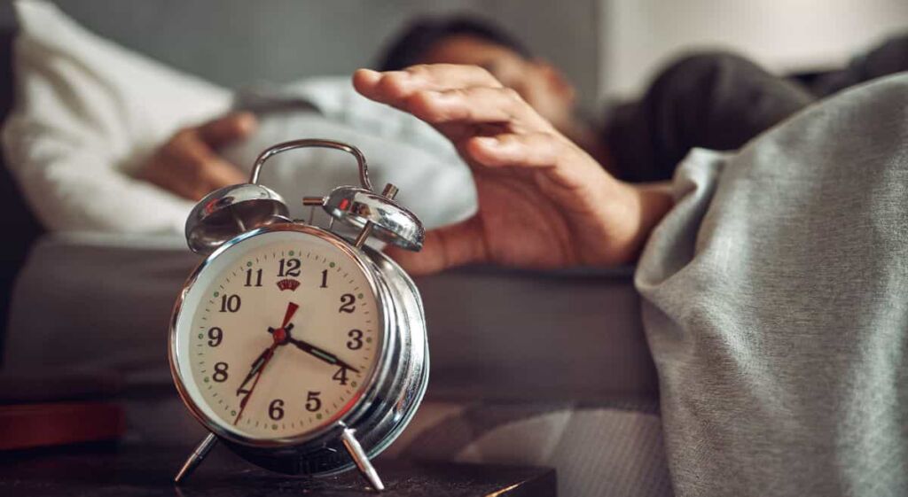 Man in bed reaching for a silver double bell alarm clock.