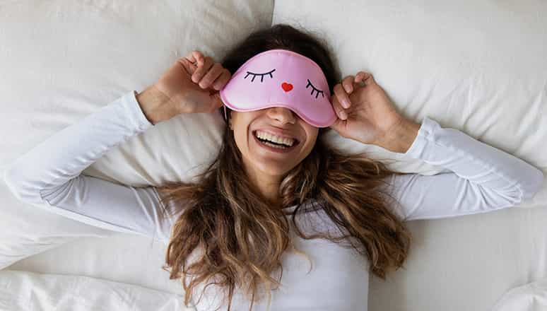 Laughing woman laying in bed wearing a pink eye mask with closed eyelid icons on it.